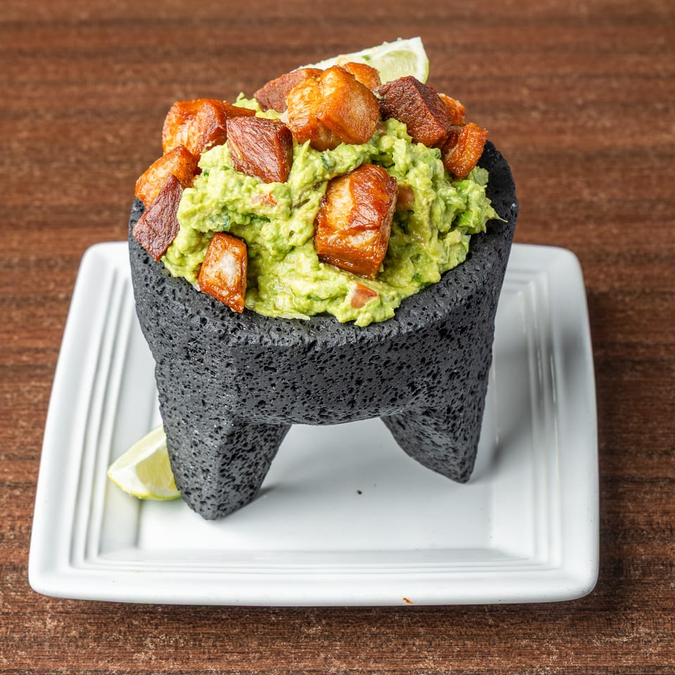 Guacamole with crispy pork belly chunks and lime wedges, served in a dark stone molcajete.