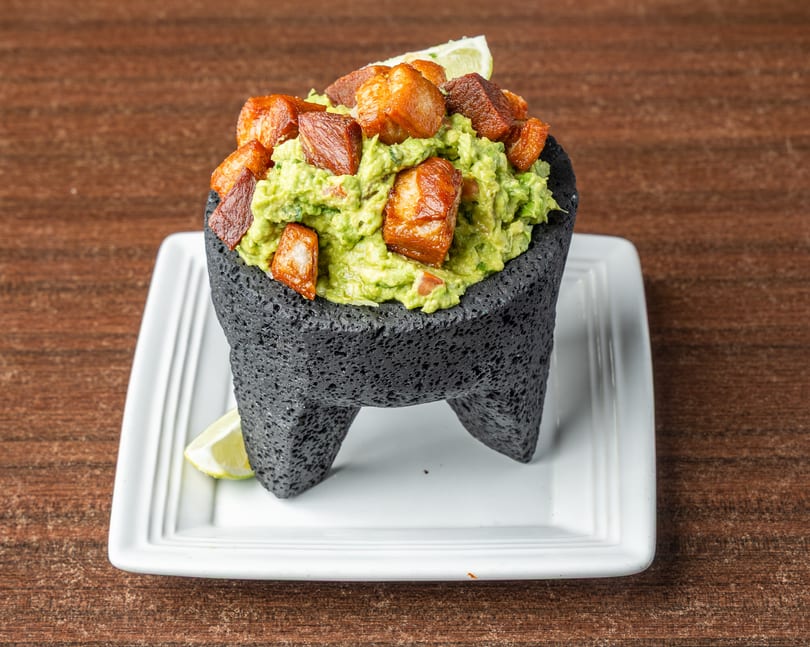 Guacamole with crispy pork belly chunks and lime wedges, served in a dark stone molcajete.