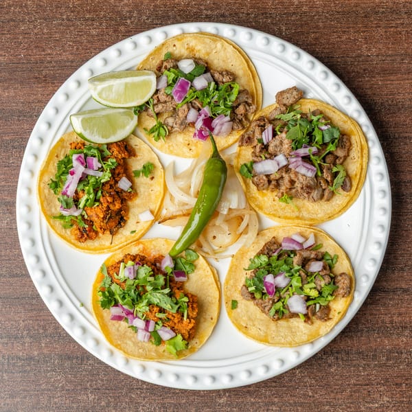 Five street tacos with various fillings, cilantro, red onion, lime wedges, and a green chili.