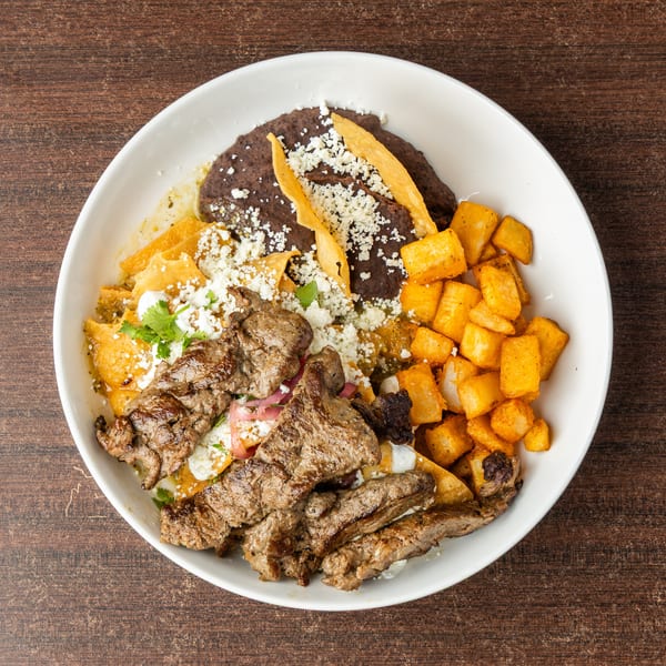 A white bowl filled with steak, chilaquiles, black beans, and fried potatoes on a wooden table.
