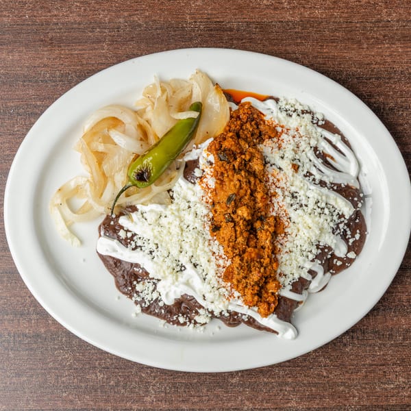 A white plate holds a Mexican dish with dark sauce, crumbled cheese, chorizo, onions, and a green chili.