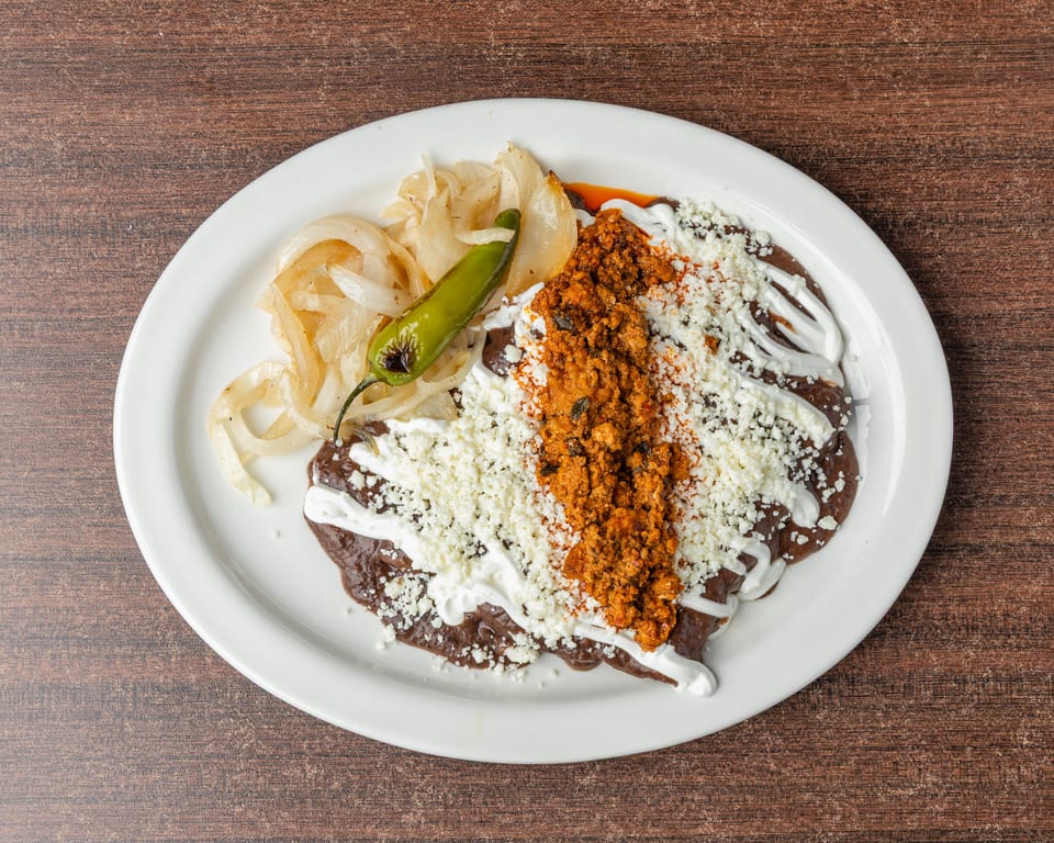 A white plate holds a Mexican dish with dark sauce, crumbled cheese, chorizo, onions, and a green chili.