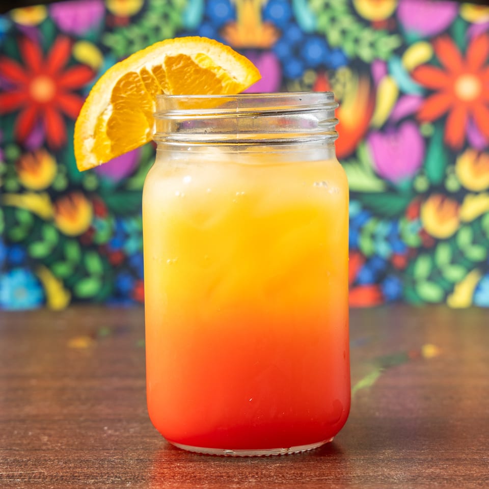 Sunrise cocktail in a mason jar with an orange slice, on a wooden table with a colorful floral background.