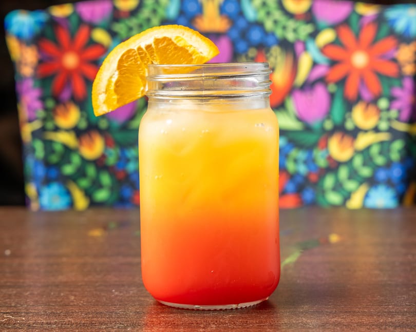 Sunrise cocktail in a mason jar with an orange slice, on a wooden table with a colorful floral background.