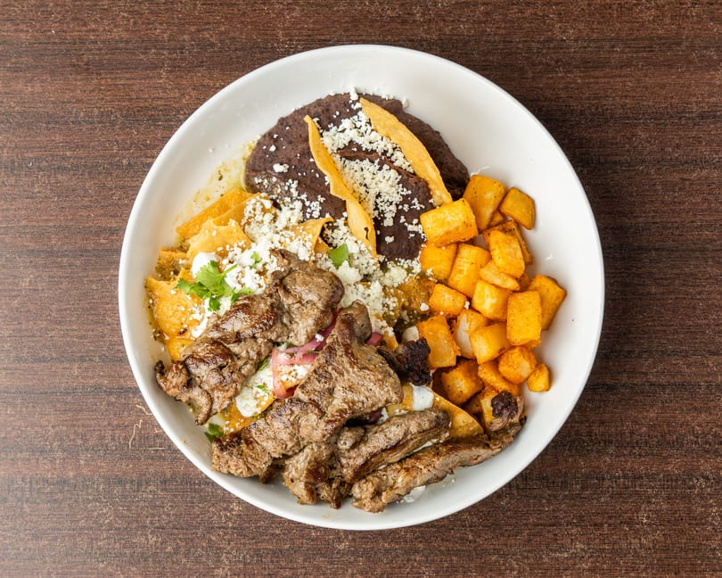 A white bowl filled with steak, chilaquiles, black beans, and diced potatoes on a wooden table.