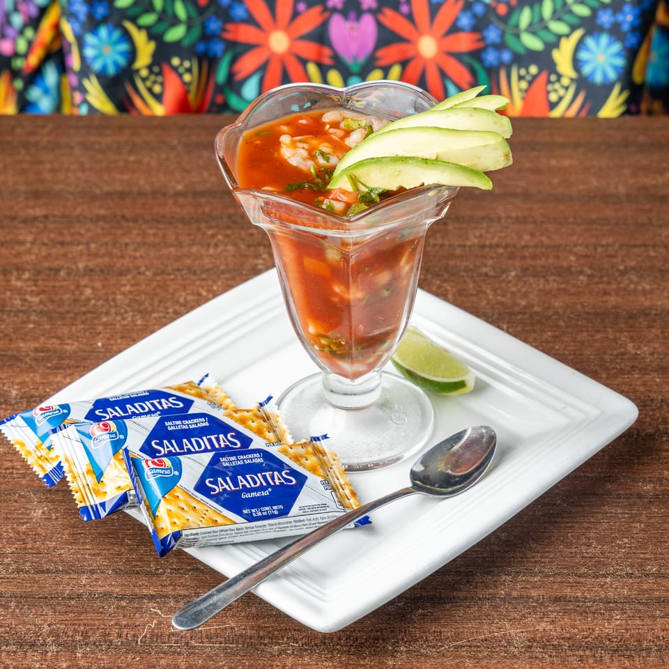 Shrimp cocktail in a glass with avocado, lime, spoon, and Saladitas crackers on a white plate.
