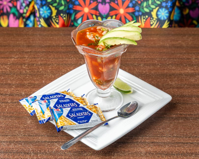 Shrimp cocktail in a glass with avocado, lime, spoon, and Saladitas crackers on a white plate.
