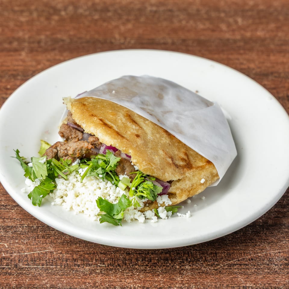 Arepa filled with meat, red onions, cilantro, and crumbled white cheese on a white plate.