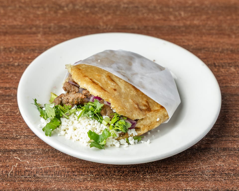 Arepa filled with meat, red onions, cilantro, and crumbled white cheese on a white plate.