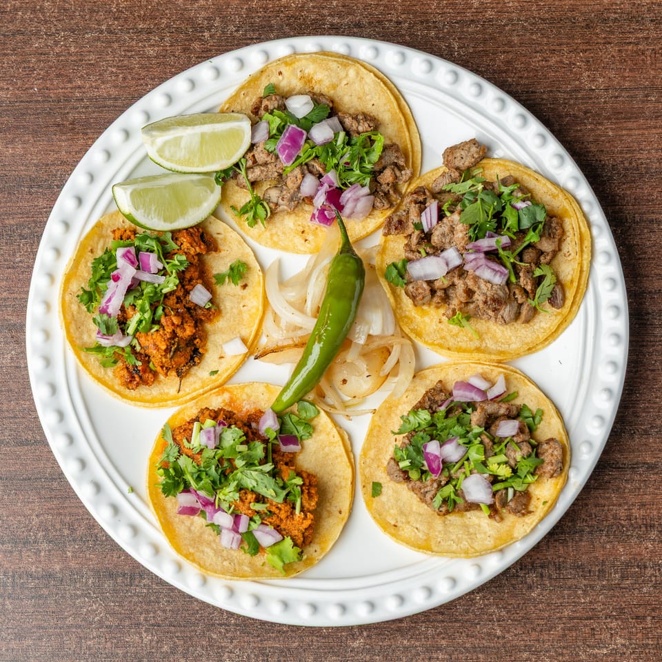 Five street tacos with various fillings, cilantro, red onion, lime, and a green chili on a white plate.