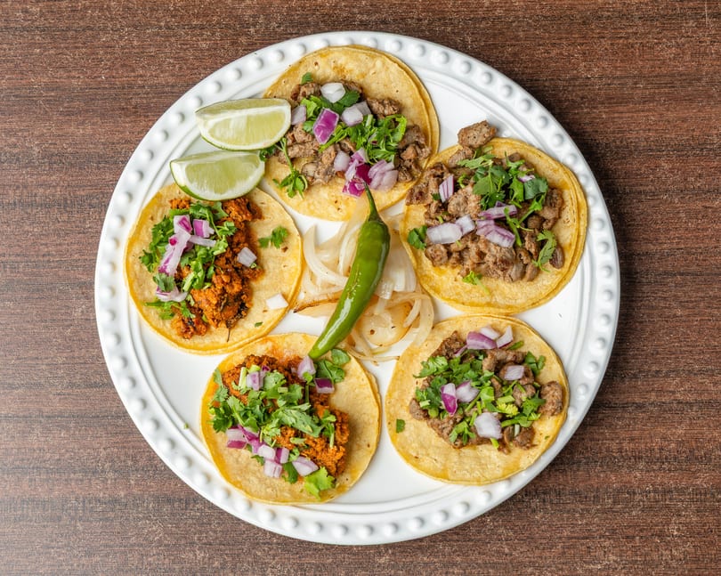 Five street tacos with various fillings, cilantro, red onion, lime, and a green chili on a white plate.